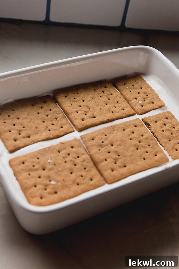 An assembled eclair cake in a clear glass baking dish, showing neat layers of golden graham crackers alternating with a pale, creamy pudding mixture, ready for the final chocolate topping.