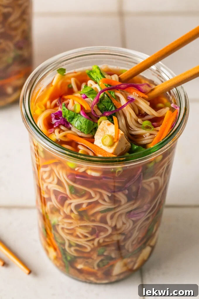 Homemade cup of noodles with ramen, veggies and broth in a glass jar with chopsticks pulling the noodles.