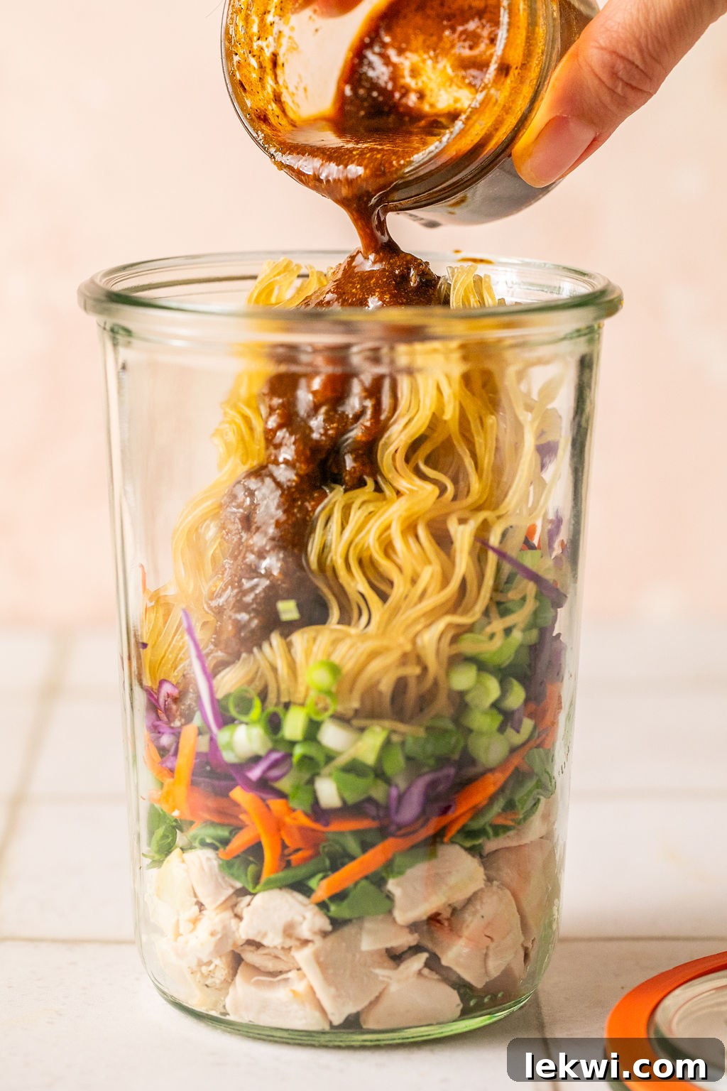 Cooked chicken, veggies, green onion, a brick of gluten free ramen layered in a glass jar with sauce pouring in.