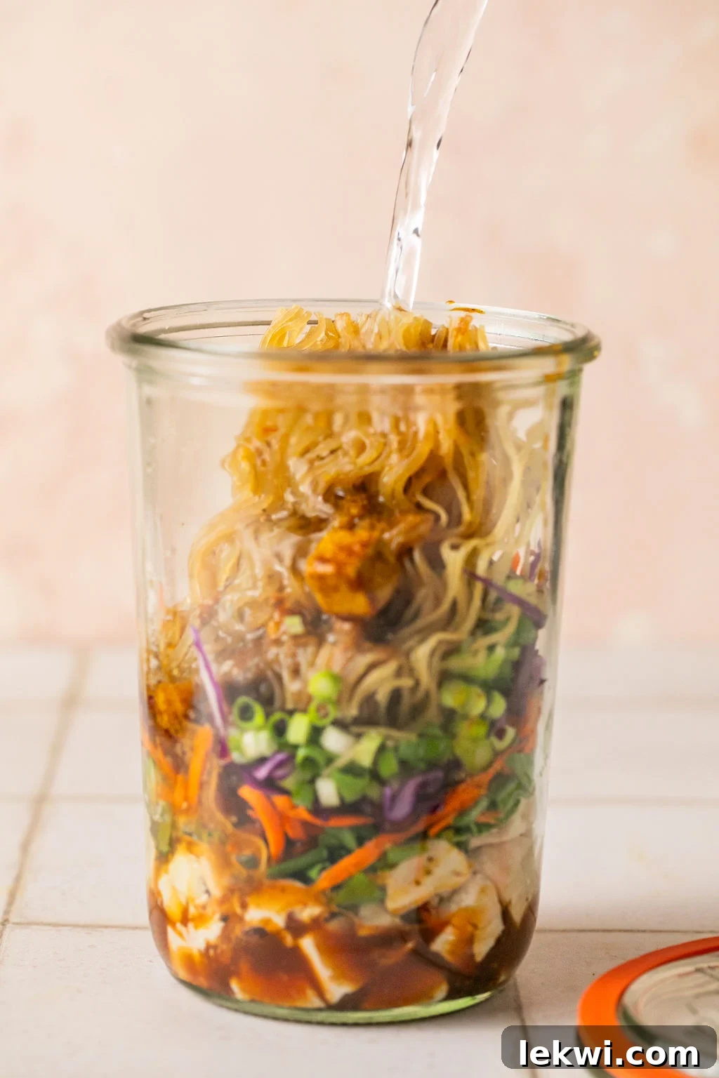 Cooked chicken, veggies, green onion, a brick of gluten free ramen layered in a glass jar with hot water pouring in.