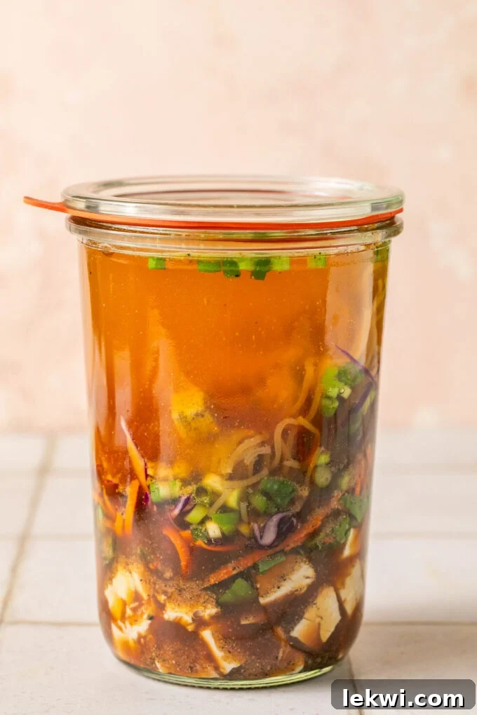 Cooked chicken, veggies, green onion, a brick of gluten free ramen layered in a glass jar with hot water.