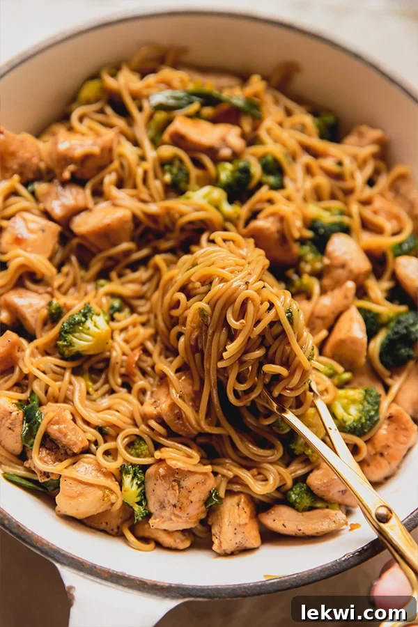 A vibrant chicken and broccoli ramen stir fry being tossed with tongs in a pan, noodles coated in sauce.