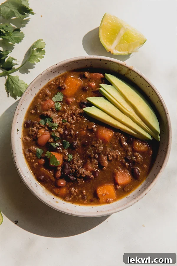 A steaming bowl of nightshade-free chili, generously topped with creamy sliced avocado and fresh cilantro, set against a rustic background, highlighting its rich texture and vibrant colors. This healthy comfort food is perfect for chilly evenings.