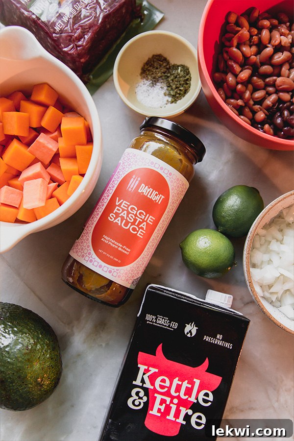 An overhead photo displaying all the fresh ingredients laid out for making nightshade-free chili, including ground beef, canned black and pinto beans, fresh limes, diced white onion, a jar of nightshade-free pasta sauce, a container of beef broth, and sliced avocado, all ready for preparation.