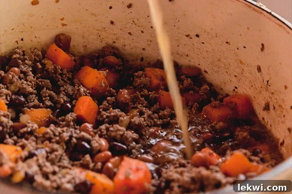 A mix of sweet potato, onion, and various beans simmering in a pot, with rich beef broth being poured in, illustrating the liquid base being added to the chili ingredients.