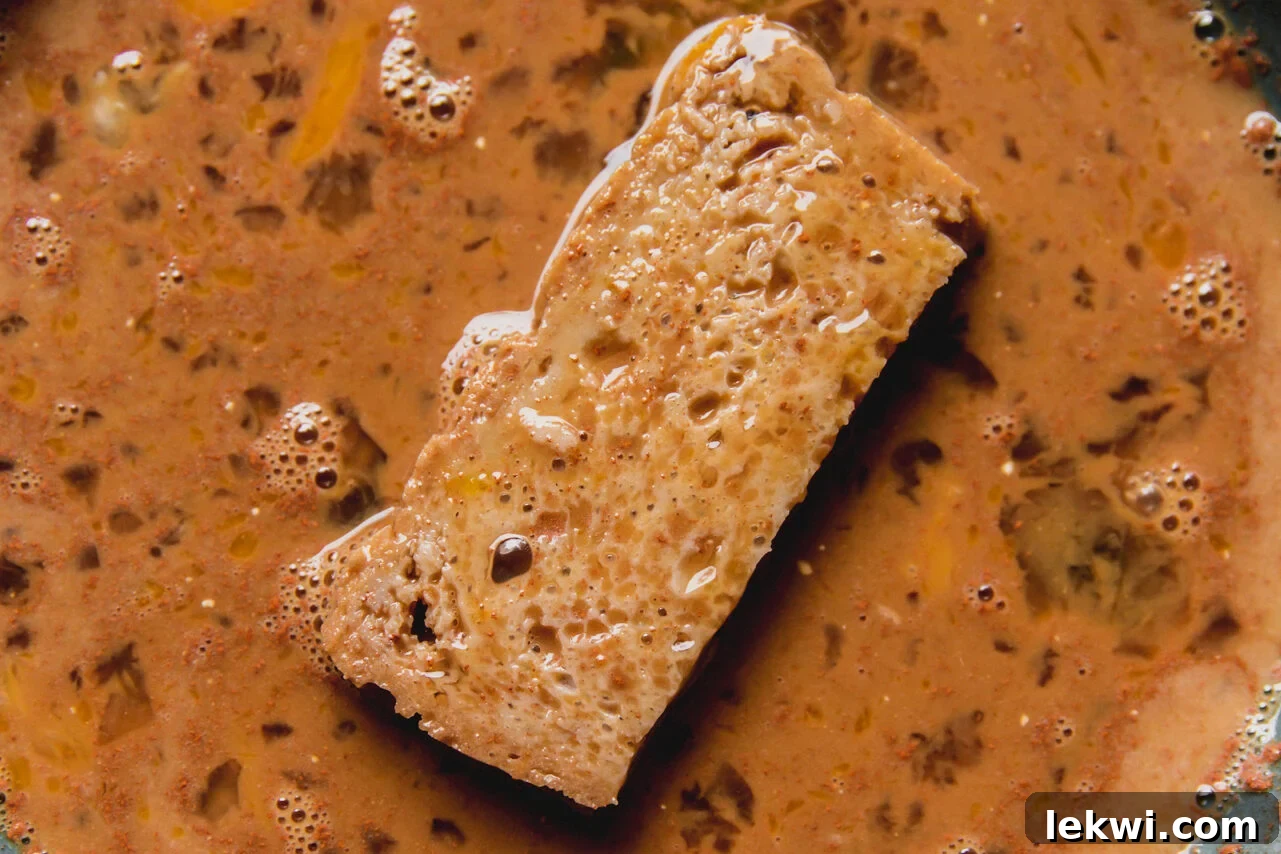 A gluten-free French toast stick being dipped into a shallow bowl filled with spiced French toast batter, ensuring even coating.