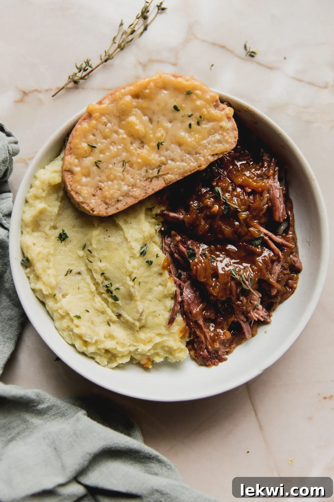 French onion pot roast in a shallow bowl with mashed sweet potato, cheese toast, and topped with herbs.