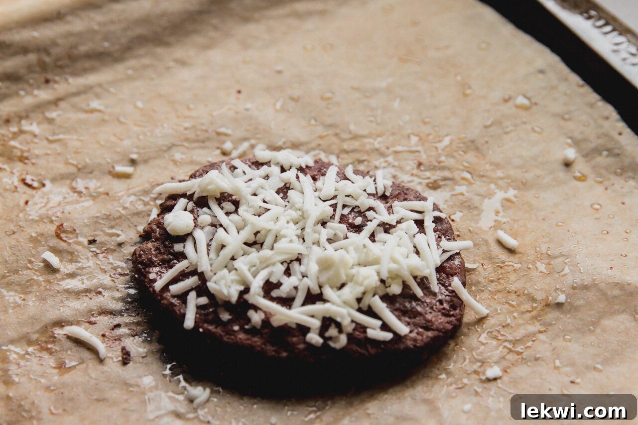 A cooked beef patty topped with a generous layer of shredded goat cheddar cheese, melted and bubbling slightly.