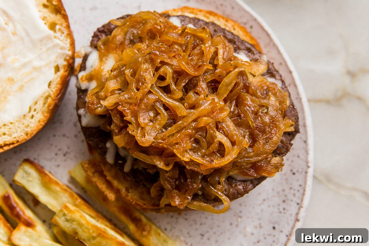 An assembled French Onion Burger, open-faced, revealing the patty, melted cheese, and a generous heap of caramelized onions on a gluten-free bun, served with fries.