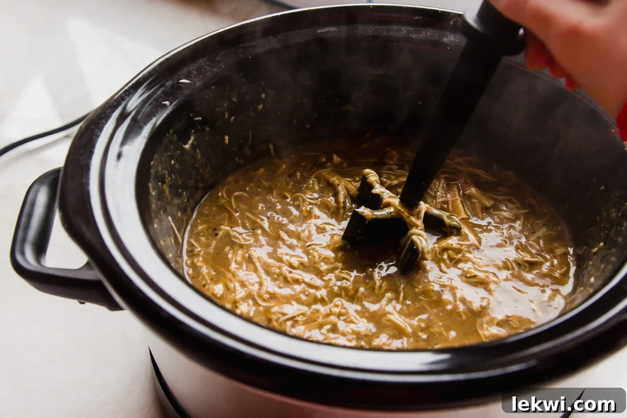 Cooked chicken being shredded into the creamy sauce within the slow cooker.