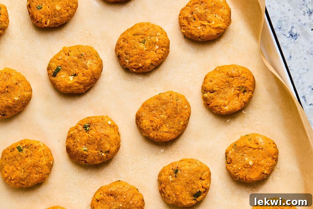 Uncooked sweet potato chicken poppers, shaped and slightly flattened, are neatly arranged on a parchment-lined baking sheet, ready for the oven.