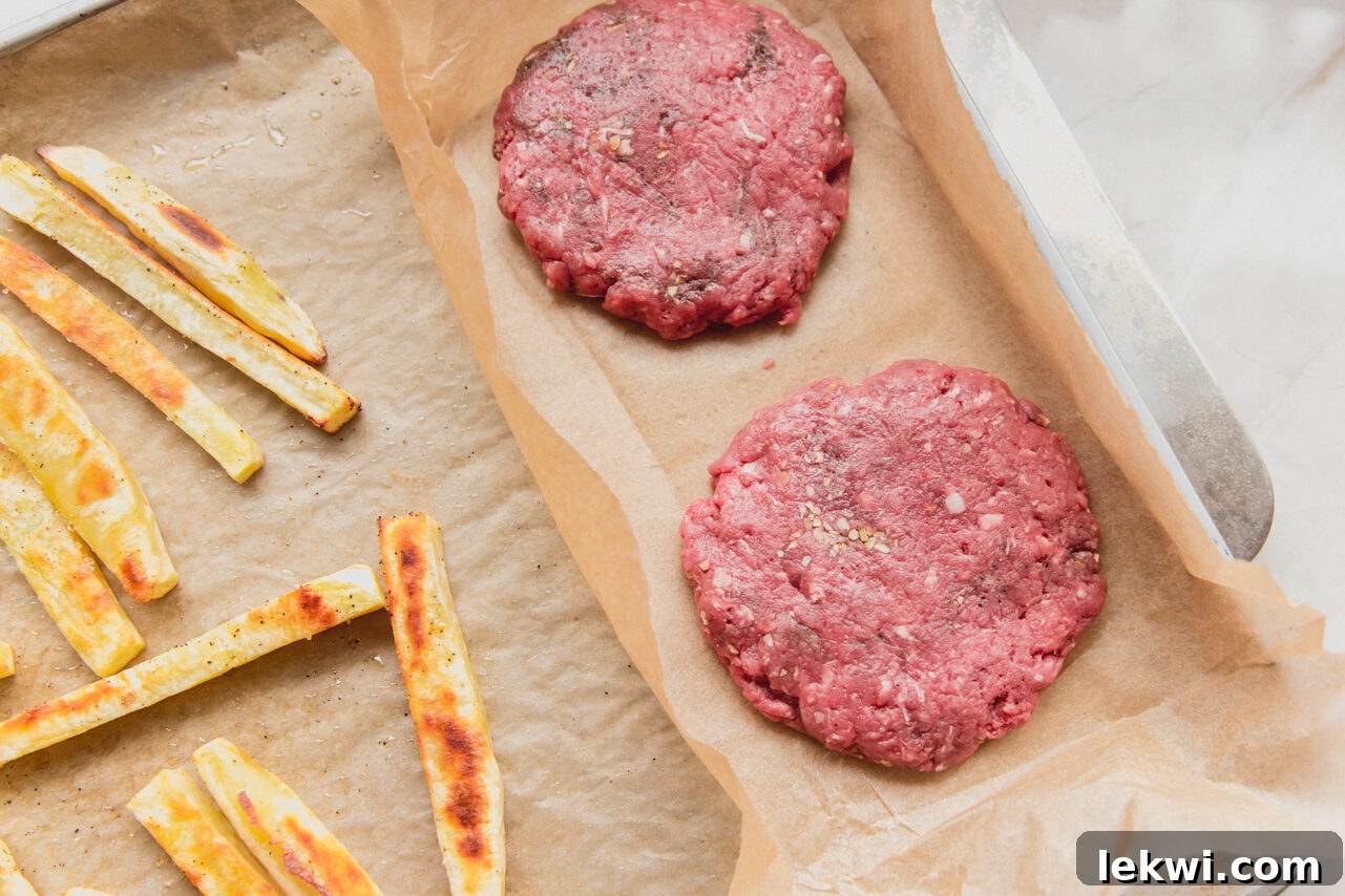A sheet pan with partially cooked fries and raw burger patties, separated by parchment paper.