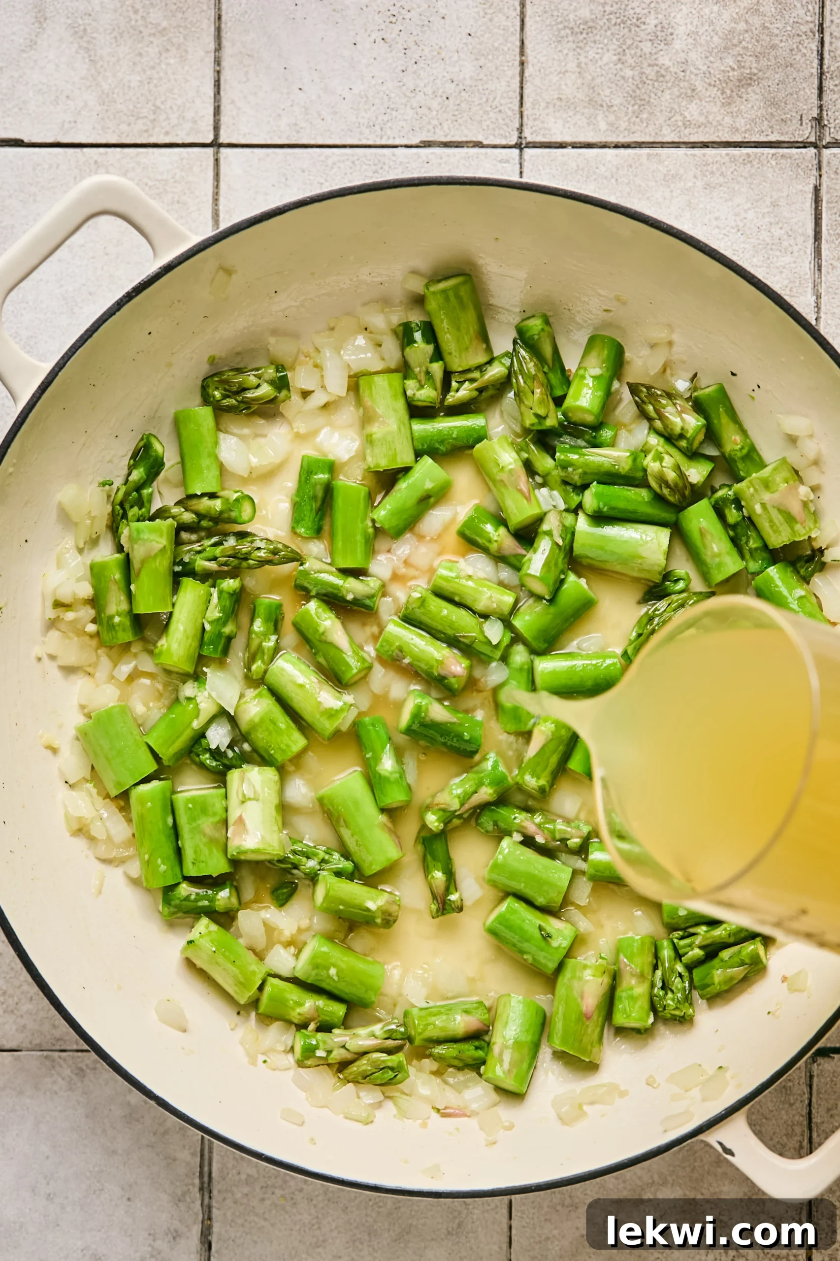 Rich chicken broth being poured into a pan, joining fragrant sautéed onion, garlic, and crisp asparagus.