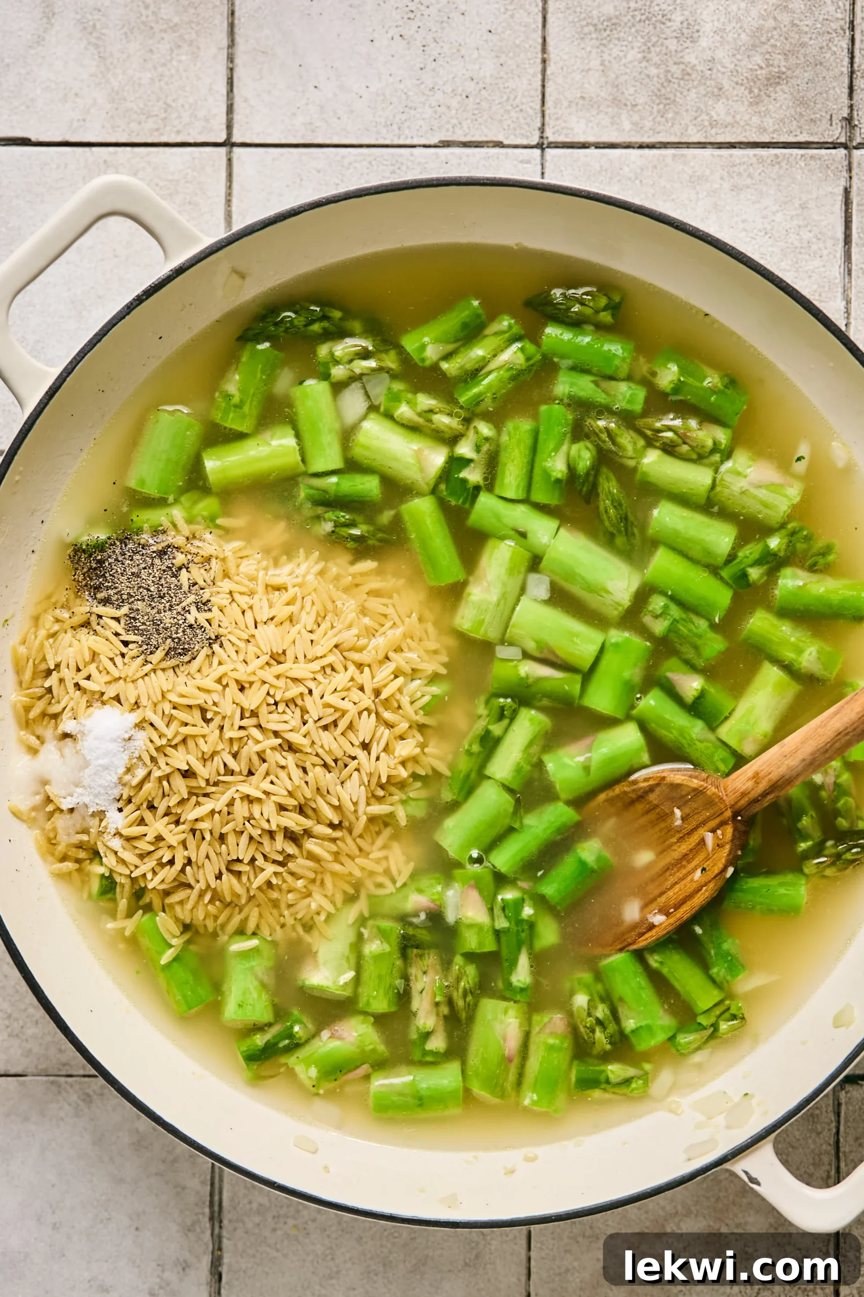 Gluten-free orzo, vibrant asparagus, and simmering broth creating a creamy base in a large cast iron pan.