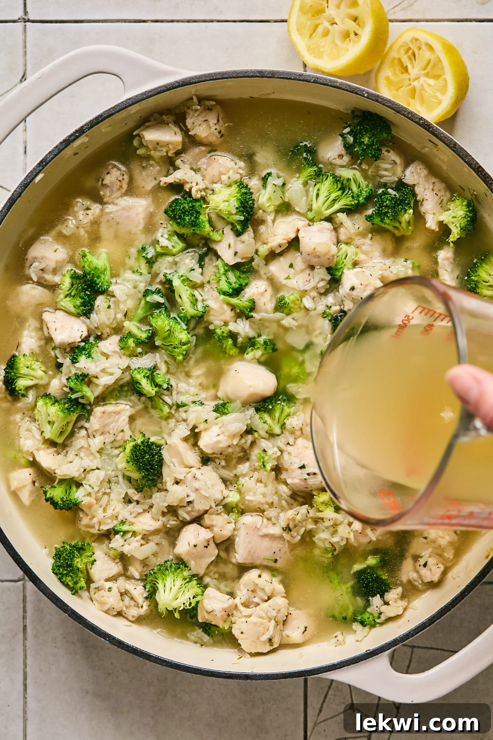 Chicken broth and lemon juice being poured into the pan with the chicken, broccoli, and rice mixture, ready to simmer.