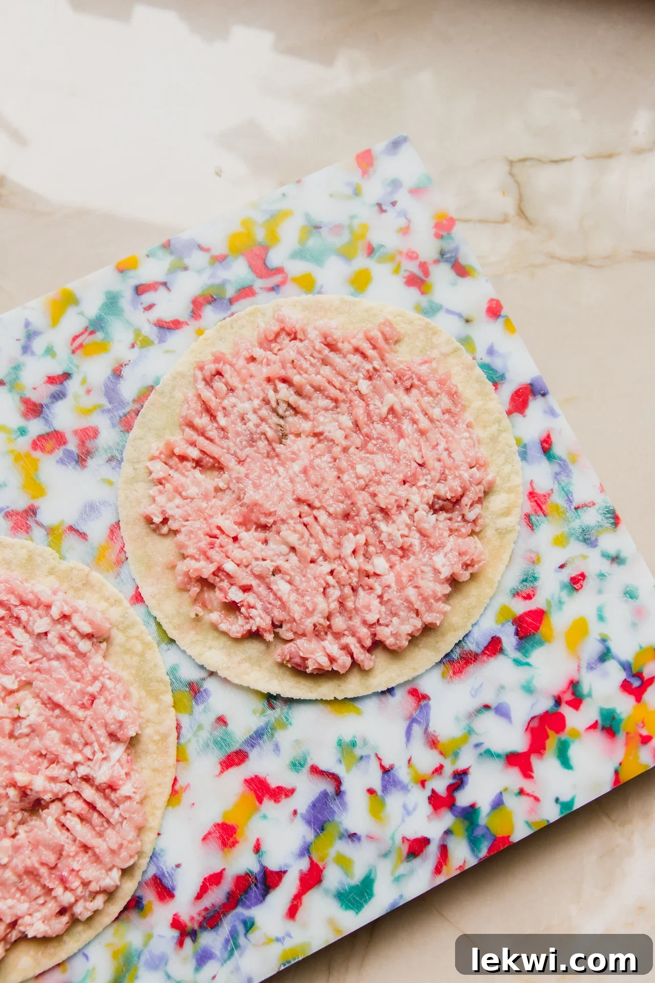 Two gluten-free tortillas laid out, each with a thin, even layer of seasoned ground chicken spread across its center.