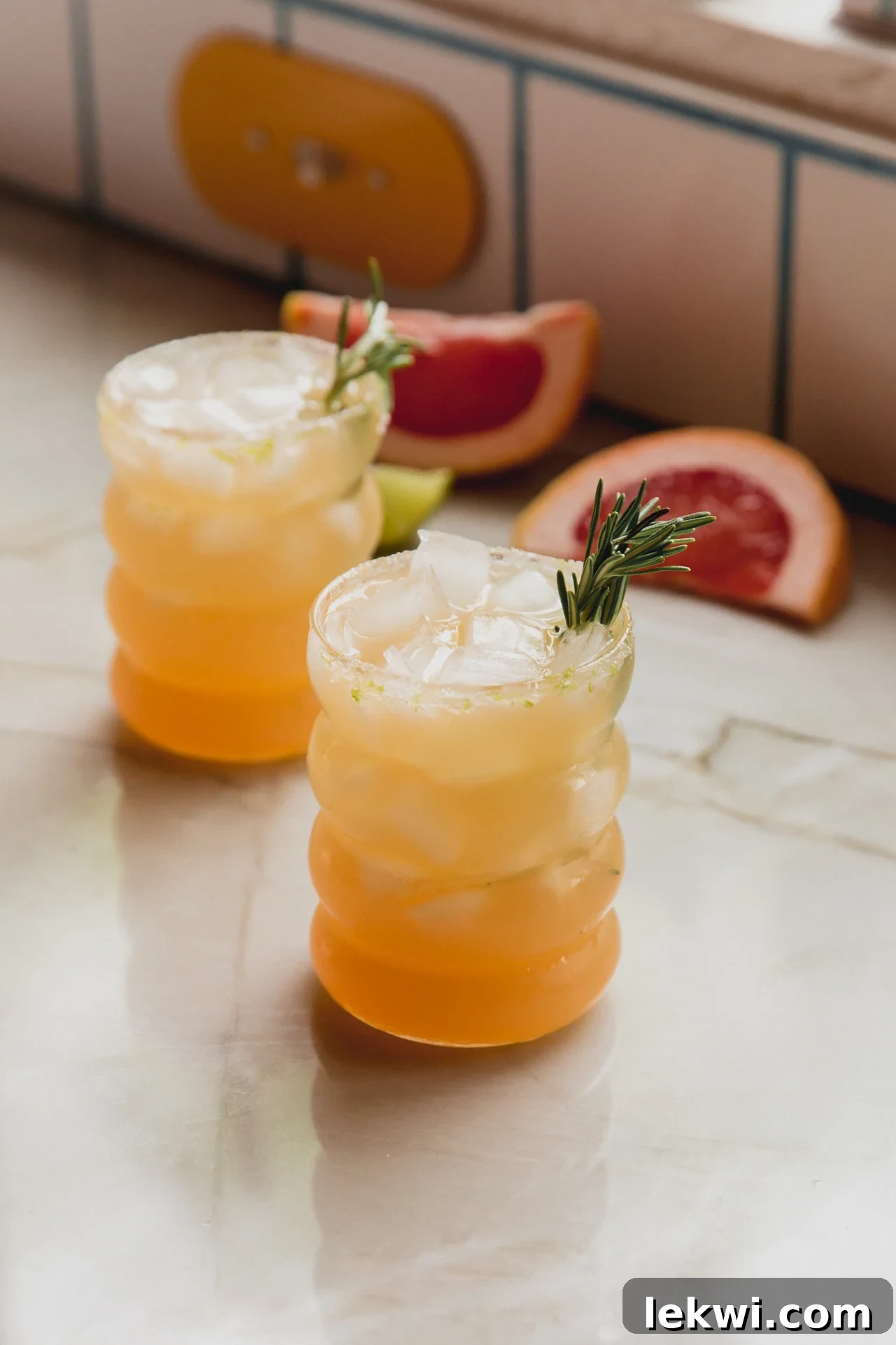 Two vibrant grapefruit paloma mocktails, garnished with fresh rosemary sprigs, standing on a rustic wooden surface.