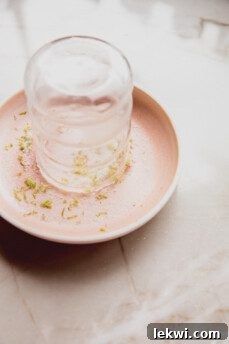 A cocktail glass being dipped into a mixture of lime zest and sweetener on a small plate to create a flavorful rim.