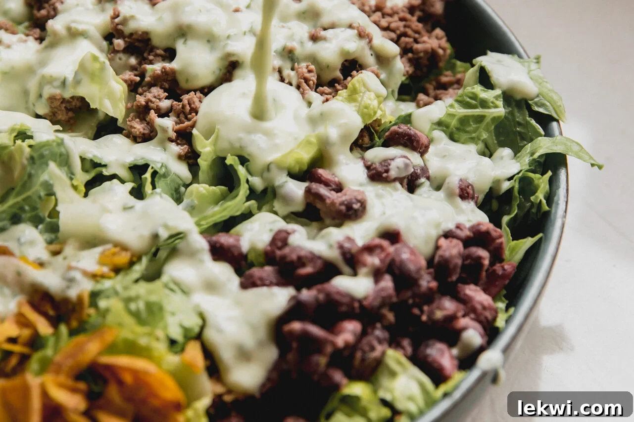 A glass jar with homemade taco salad dressing being poured generously over a colorful taco salad, topped with fresh cilantro.