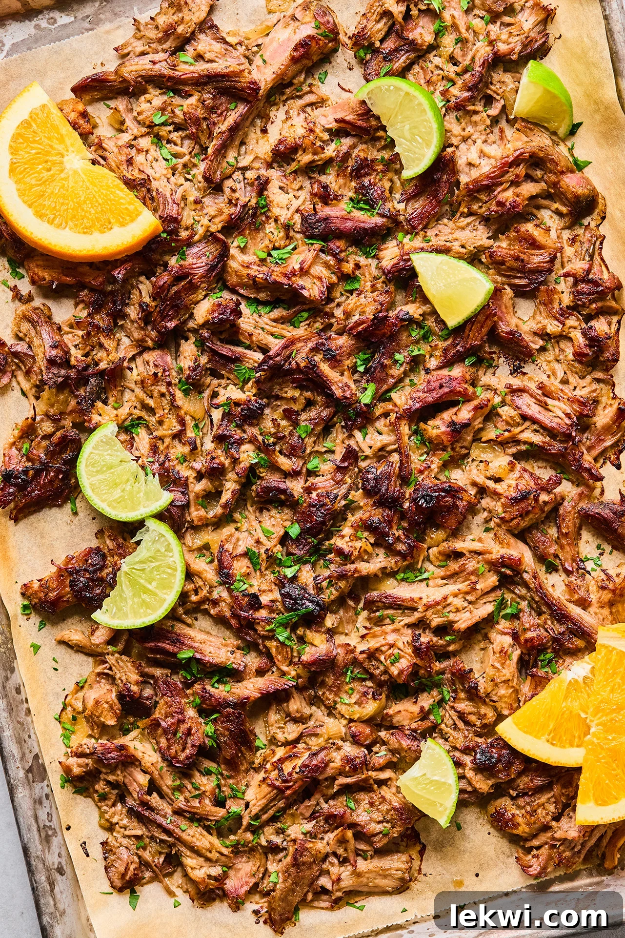 Crispy carnitas on a parchment lined baking sheet topped with chopped cilantro with lime wedges and orange wedges.