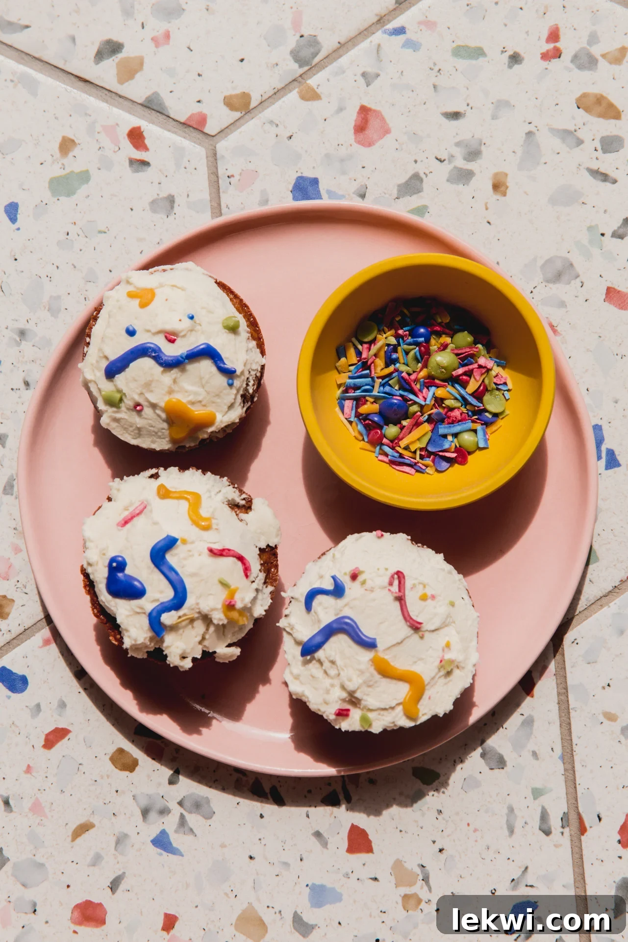 A beautifully arranged plate featuring cupcakes adorned with a mix of colorful homemade sprinkles, alongside a small bowl brimming with extra sprinkles.