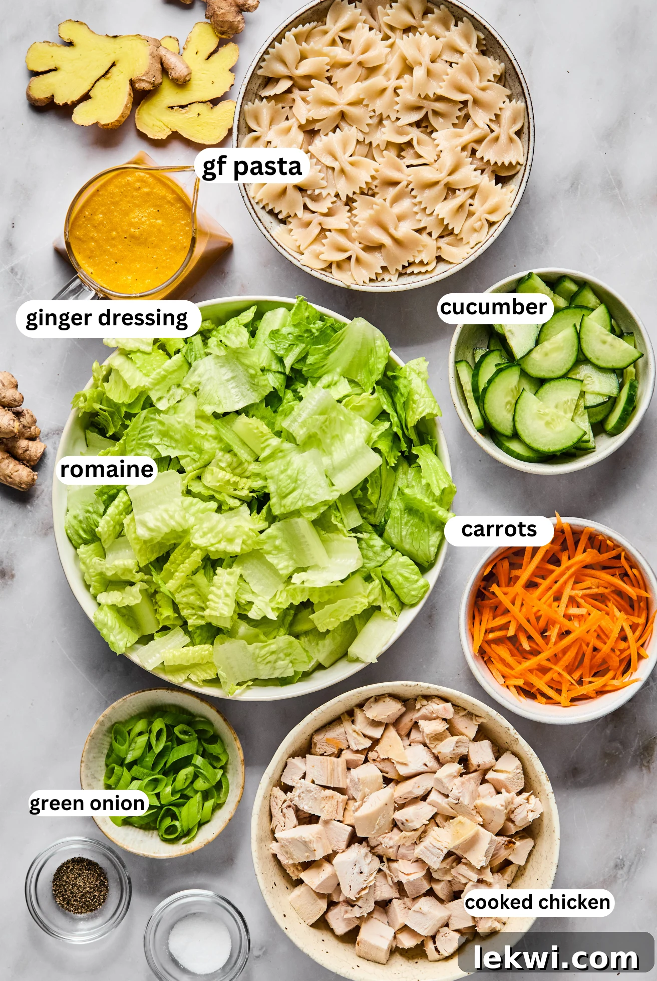 Individual bowls of Japanese Ginger Pasta Salad ingredients: gluten-free farfalle, chopped romaine, sliced cucumber, shredded carrots, cooked cubed chicken, and a jar of ginger dressing.