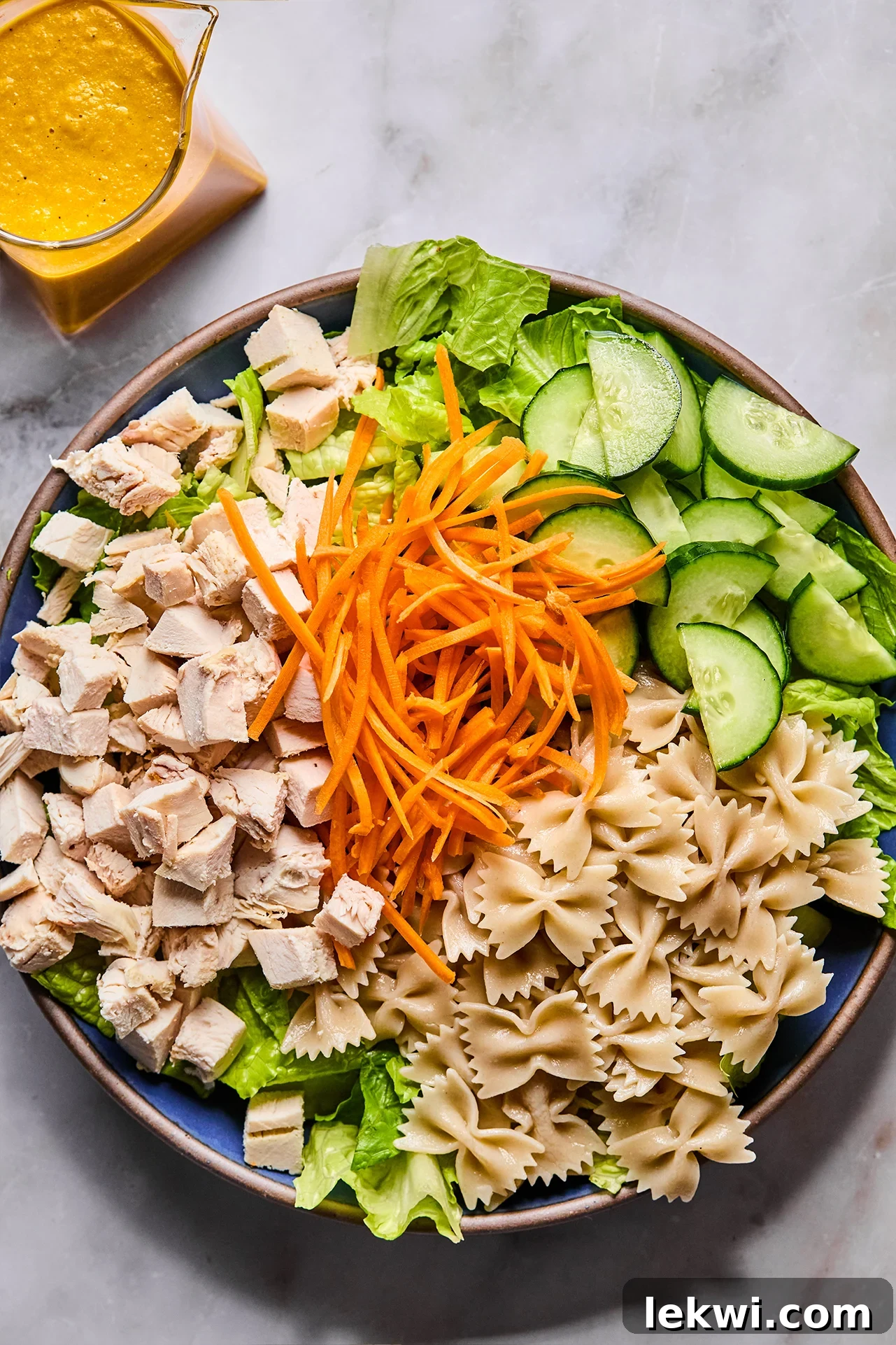 A large salad bowl filled with cooked gluten-free farfalle pasta, chopped romaine lettuce, sliced cucumber, shredded carrots, and cubed cooked chicken.