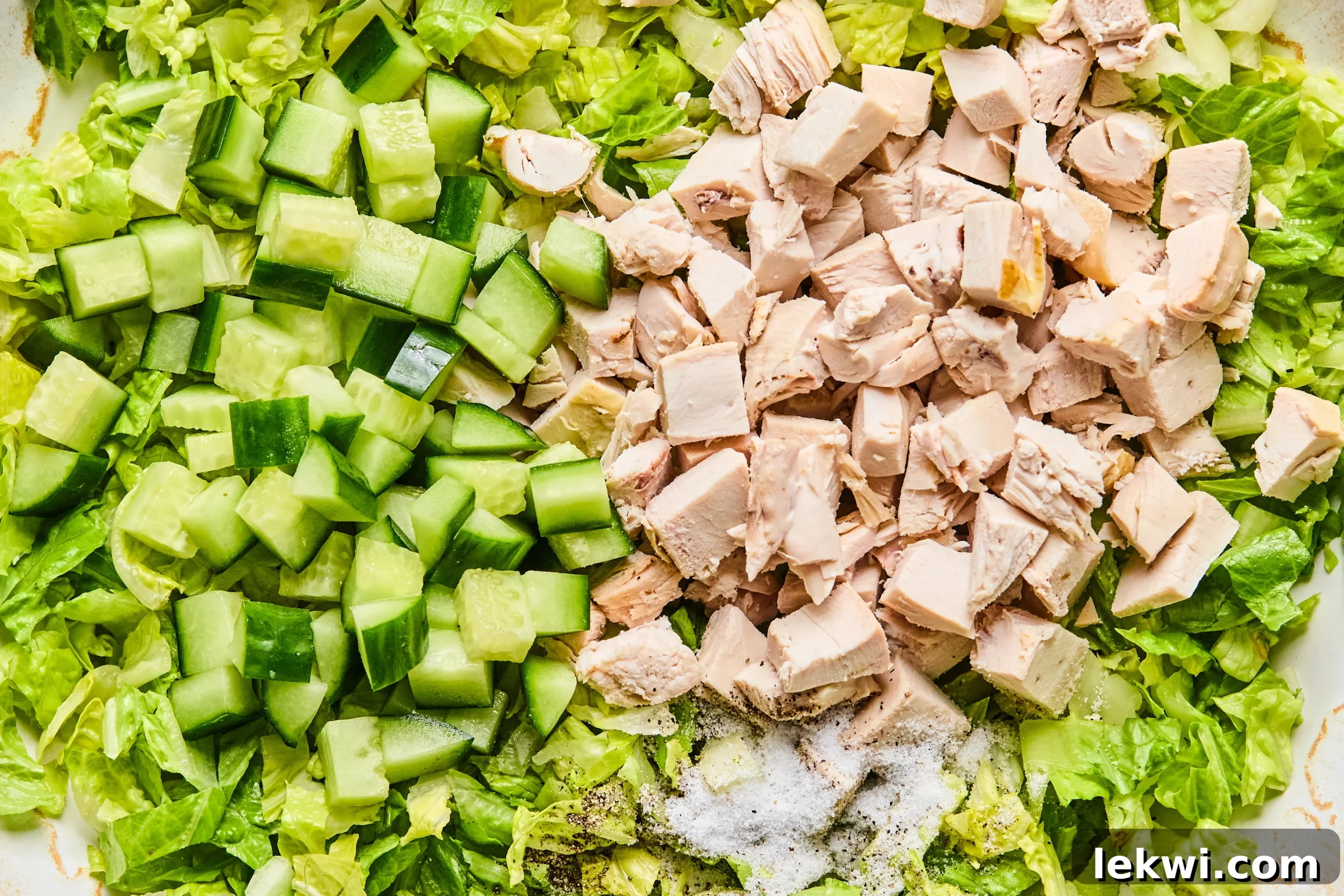 A large salad bowl filled with freshly chopped romaine lettuce, tender cooked chicken, and crisp diced cucumber, ready for seasoning and dressing application.