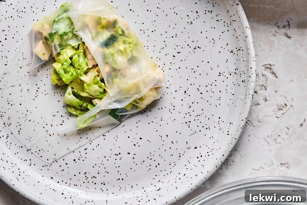 A rice paper roll actively being rolled, with the side corners neatly folded in and the bottom edge brought up and tucked over the filling.