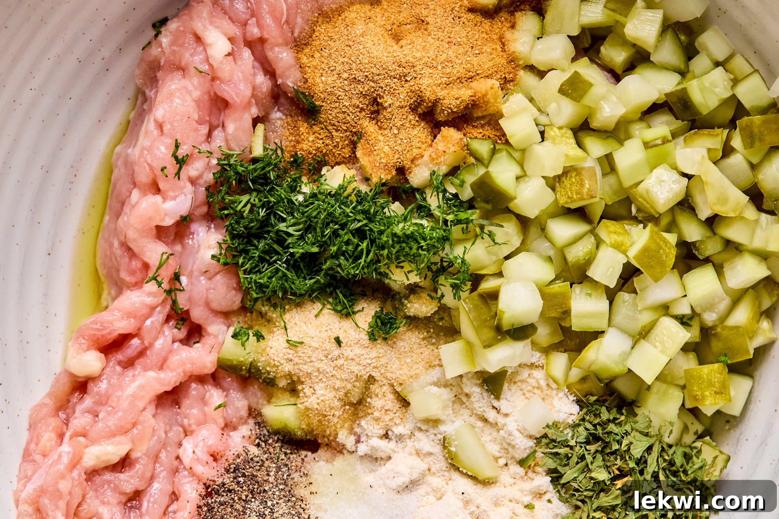 A large mixing bowl containing ground chicken, diced dill pickles, fresh dill, pickle juice, and various seasonings, unmixed.