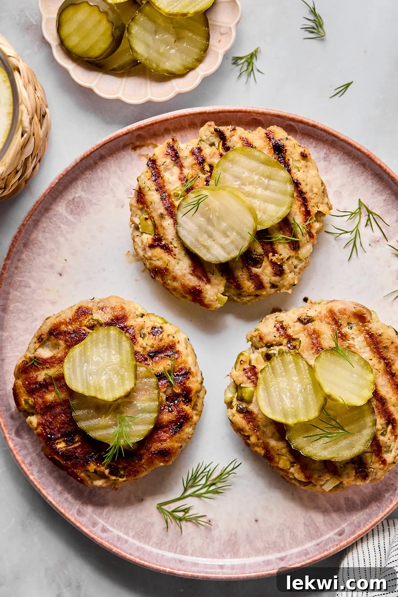 Three pickle chicken burgers topped off with sliced pickles and garnished with dill, all on a pink plate.