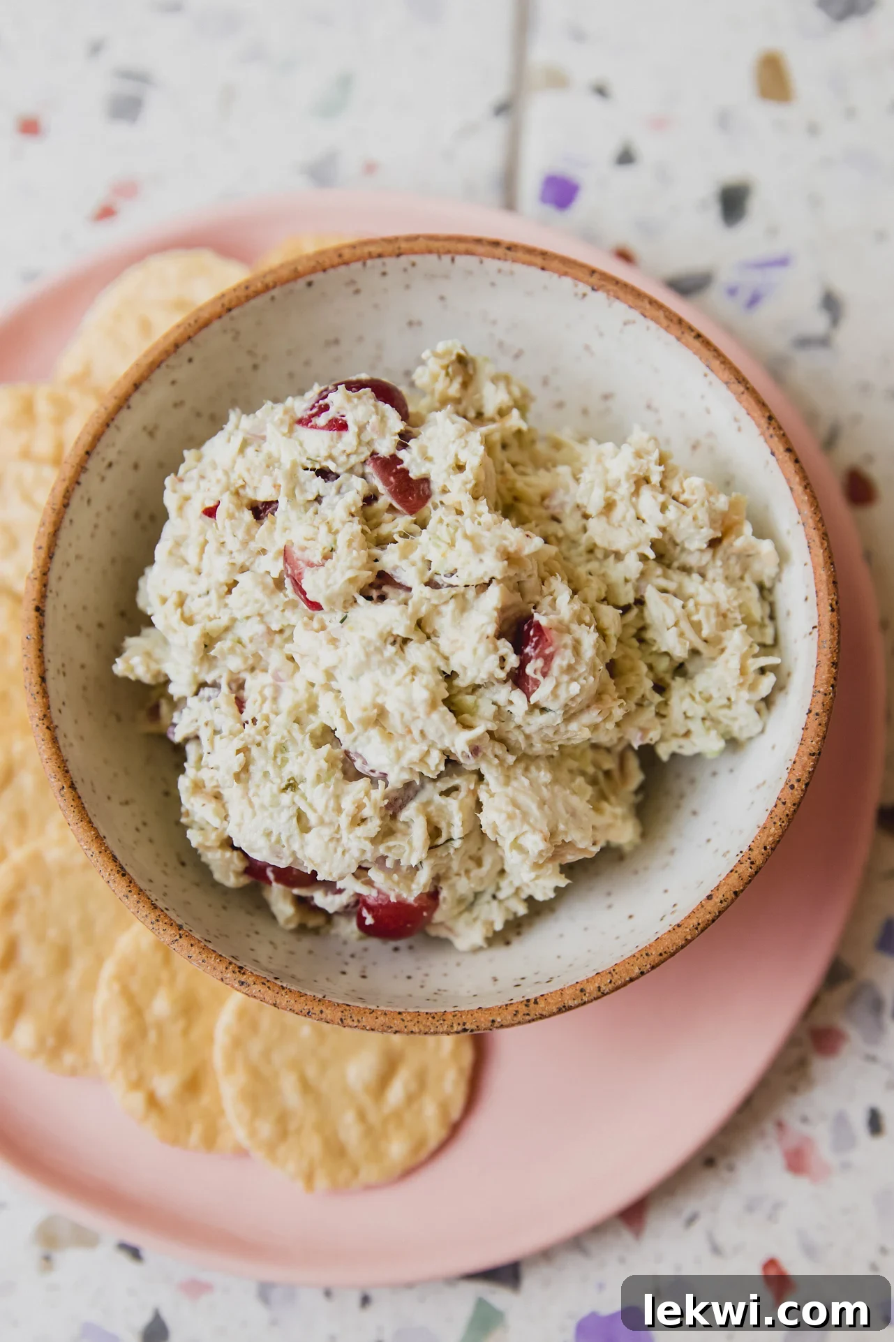 A small bowl of dairy-free grape chicken salad on a pink plate, surrounded by gluten-free crackers, showcasing a light and appetizing presentation.