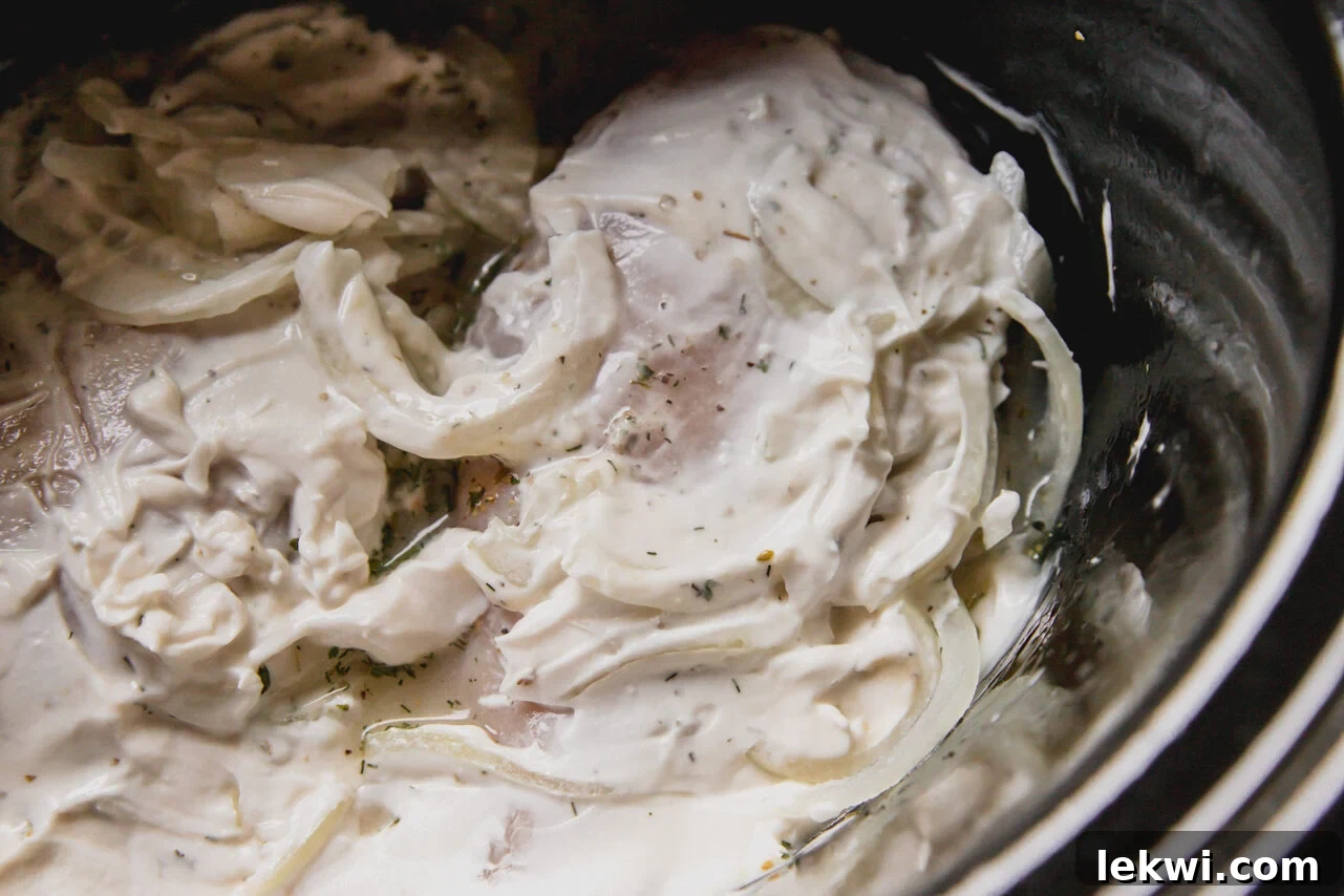 Slow cooker greek chicken uncooked in a slow cooker with sliced onions and yogurt.