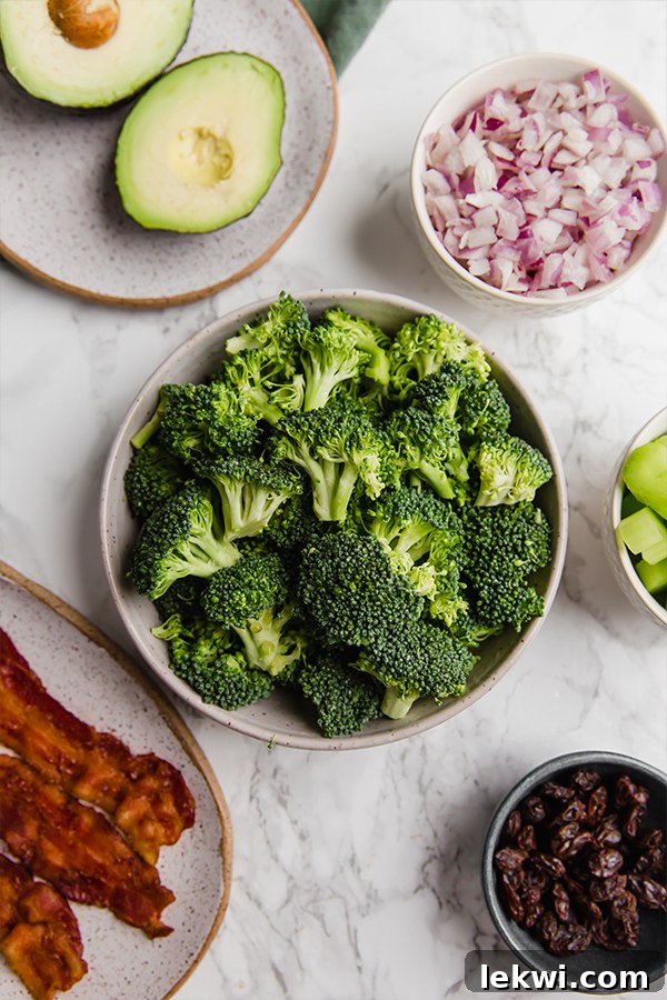 Bowls of prepped ingredients, including chopped broccoli, bacon, raisins, red onion, and celery, ready for mixing to make avocado bacon broccoli salad.