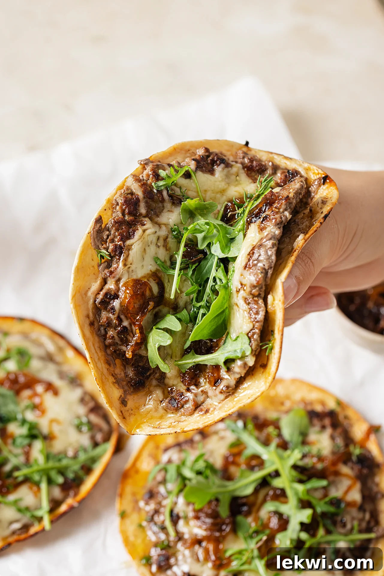 French onion smashburger tacos topped with fresh arugula, with one taco being lifted, partially folded and ready to eat.