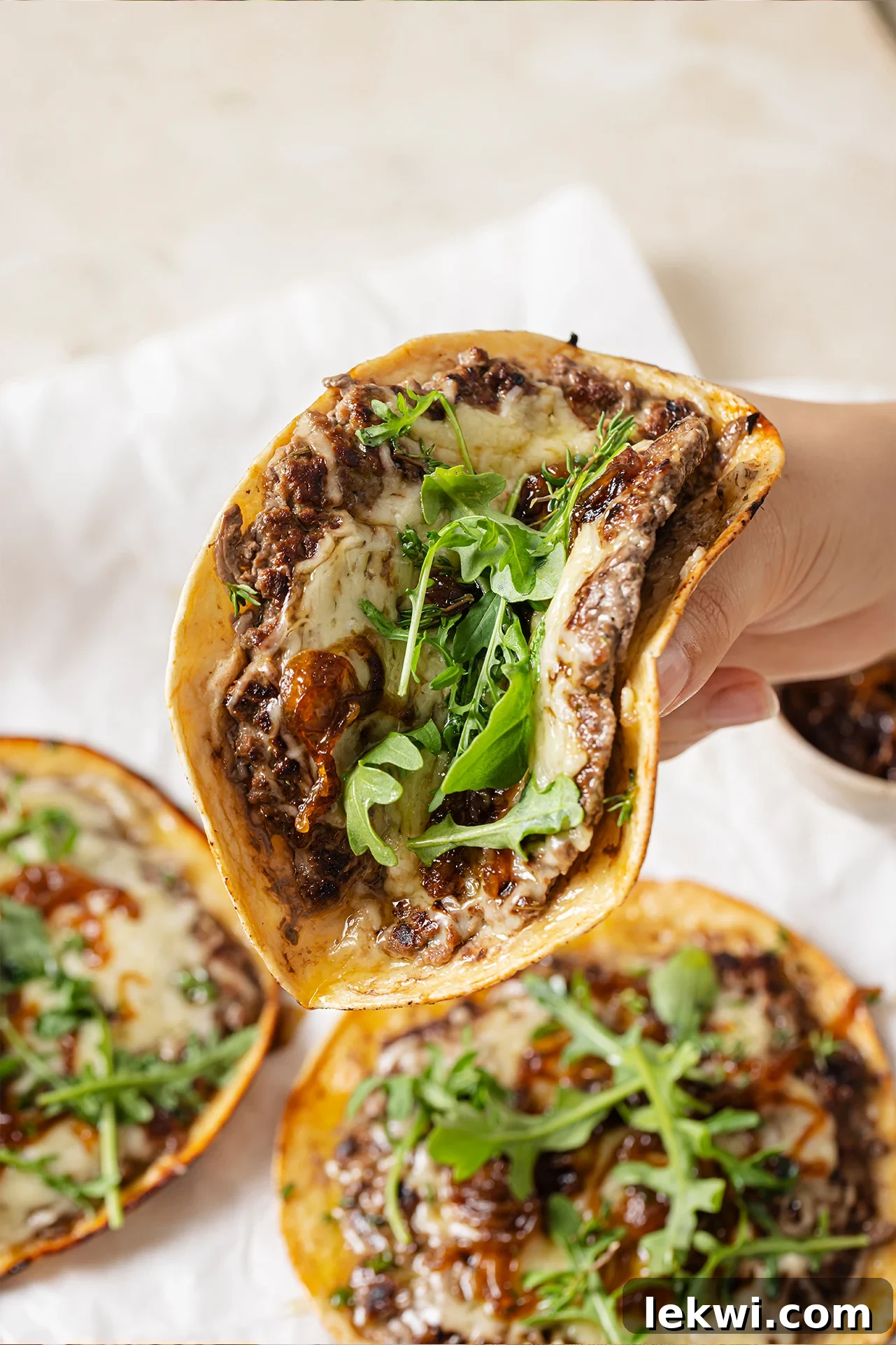 French onion smashburger tacos topped with fresh arugula, with one taco being lifted, partially folded.
