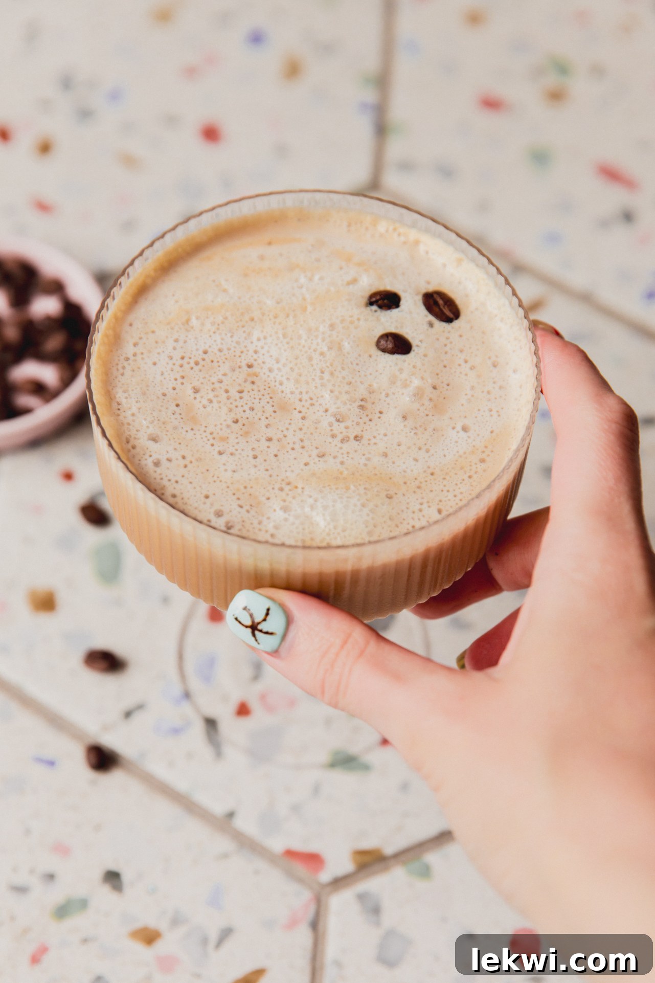 A hand holding an elegant espresso martini mocktail in a cocktail glass, garnished with espresso beans, against a soft, blurred background.