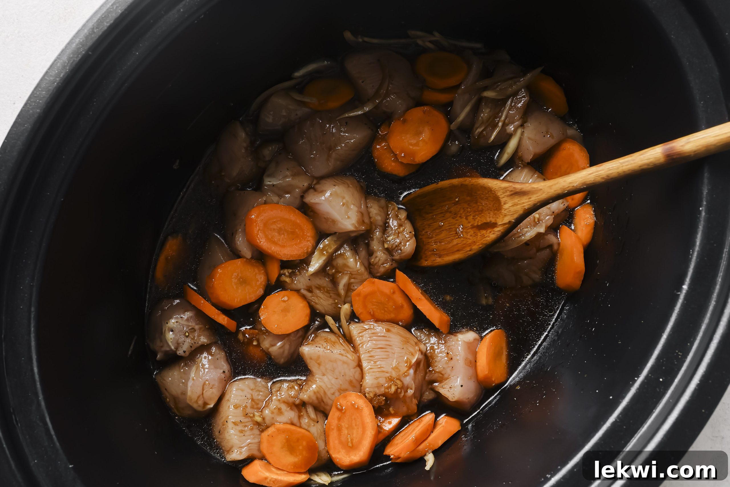 Cubed chicken and sliced carrots and onions placed in the slow cooker, with the whisked sauce poured over them, ready for cooking.