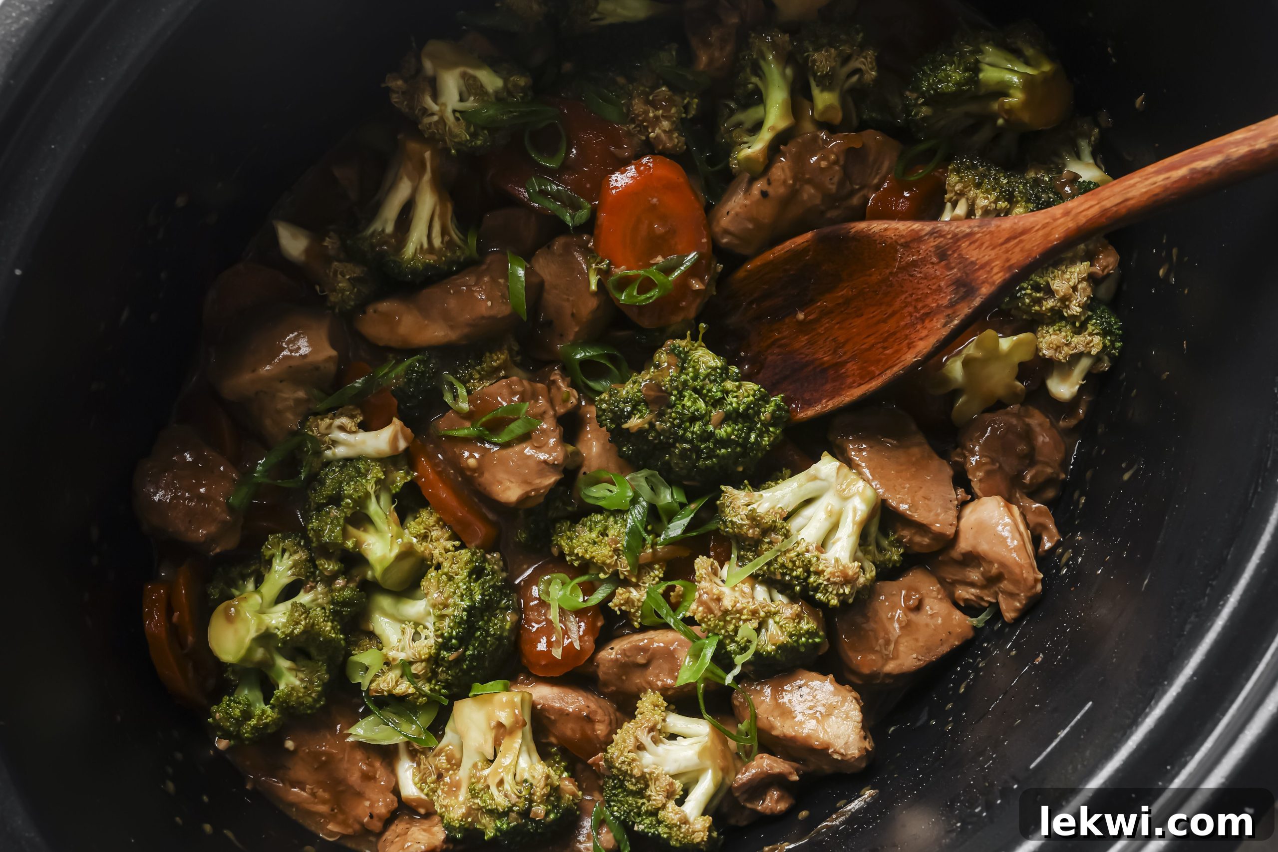 The fully cooked slow cooker chicken and broccoli, with a thick, glossy sauce, topped with vibrant green onions and a wooden spoon resting in the pot.