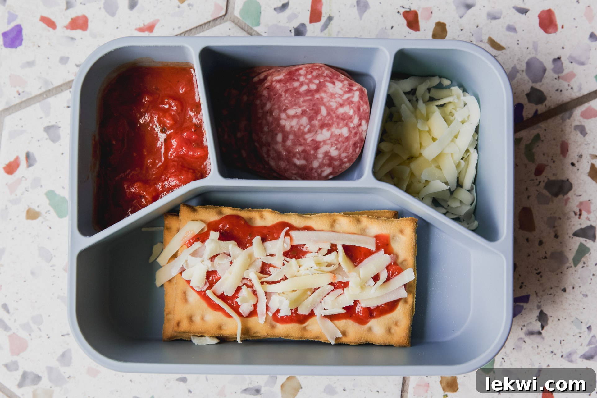 Blue lunch container with pizza sauce, meat, cheese, and a prepared flatbread pizza.