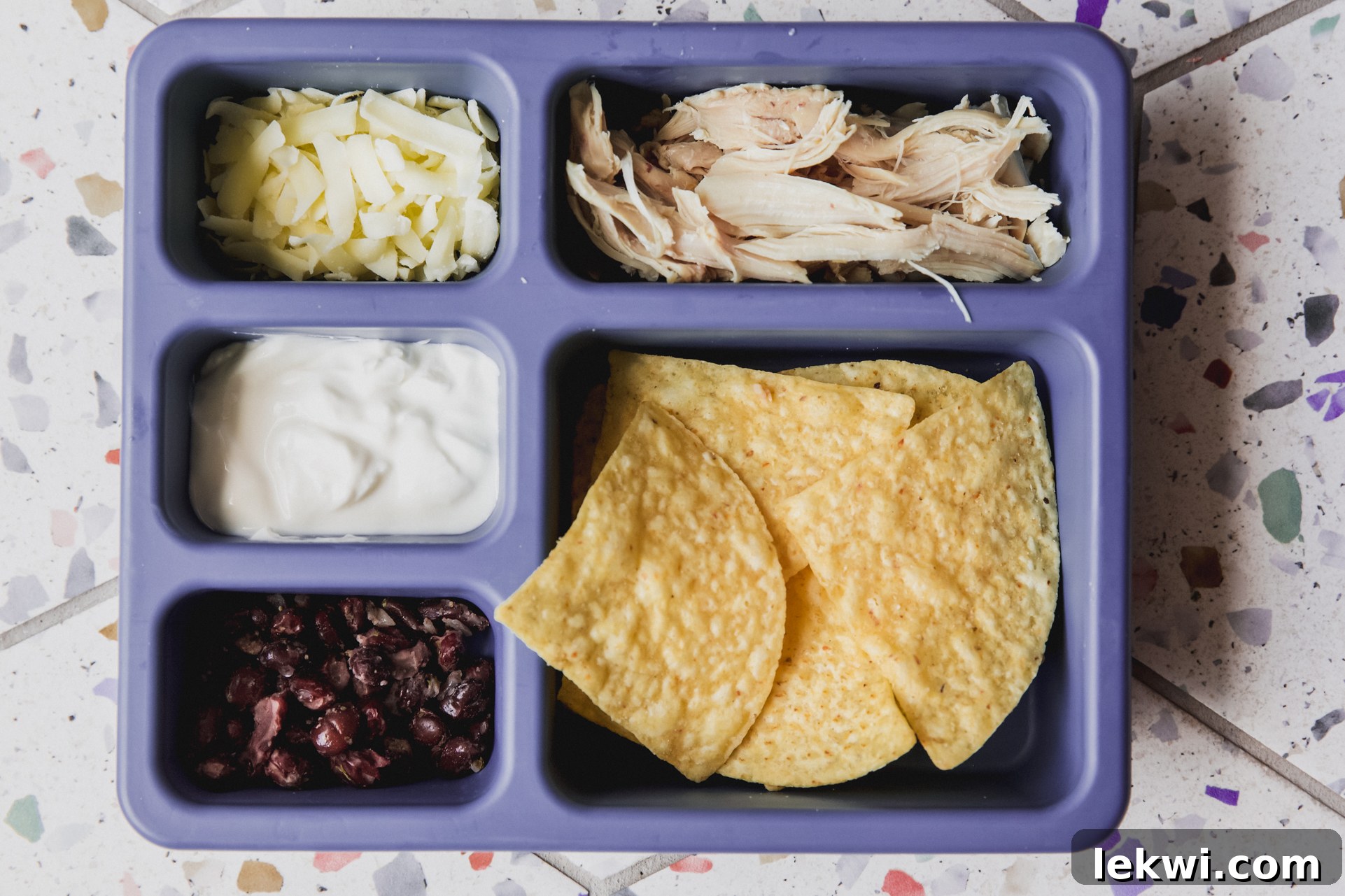 Divided lunch container with tortilla chips, shredded chicken, shredded cheese, cooked black beans and sour cream. 