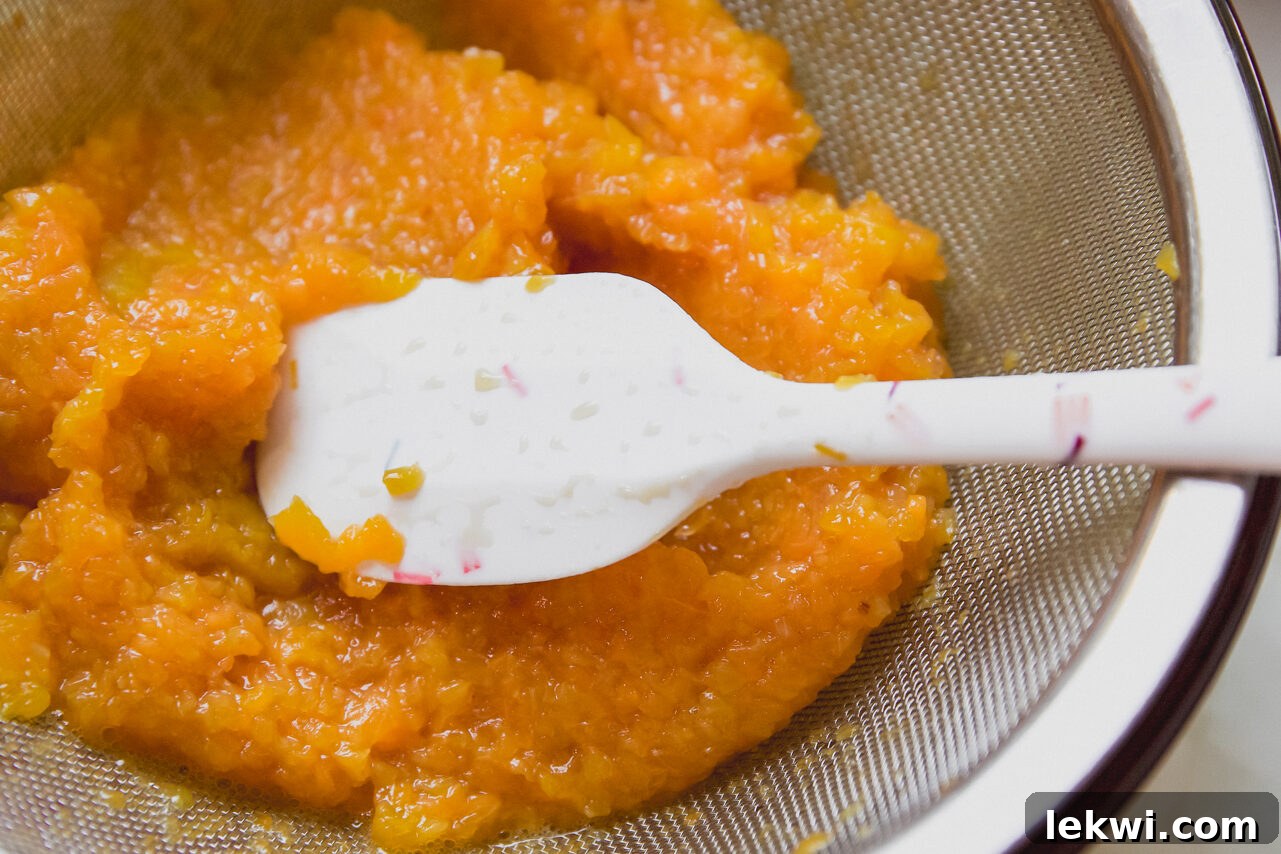 A fine mesh strainer is actively used to separate smooth peach juice from its pulp, with a silicone spatula assisting the process.