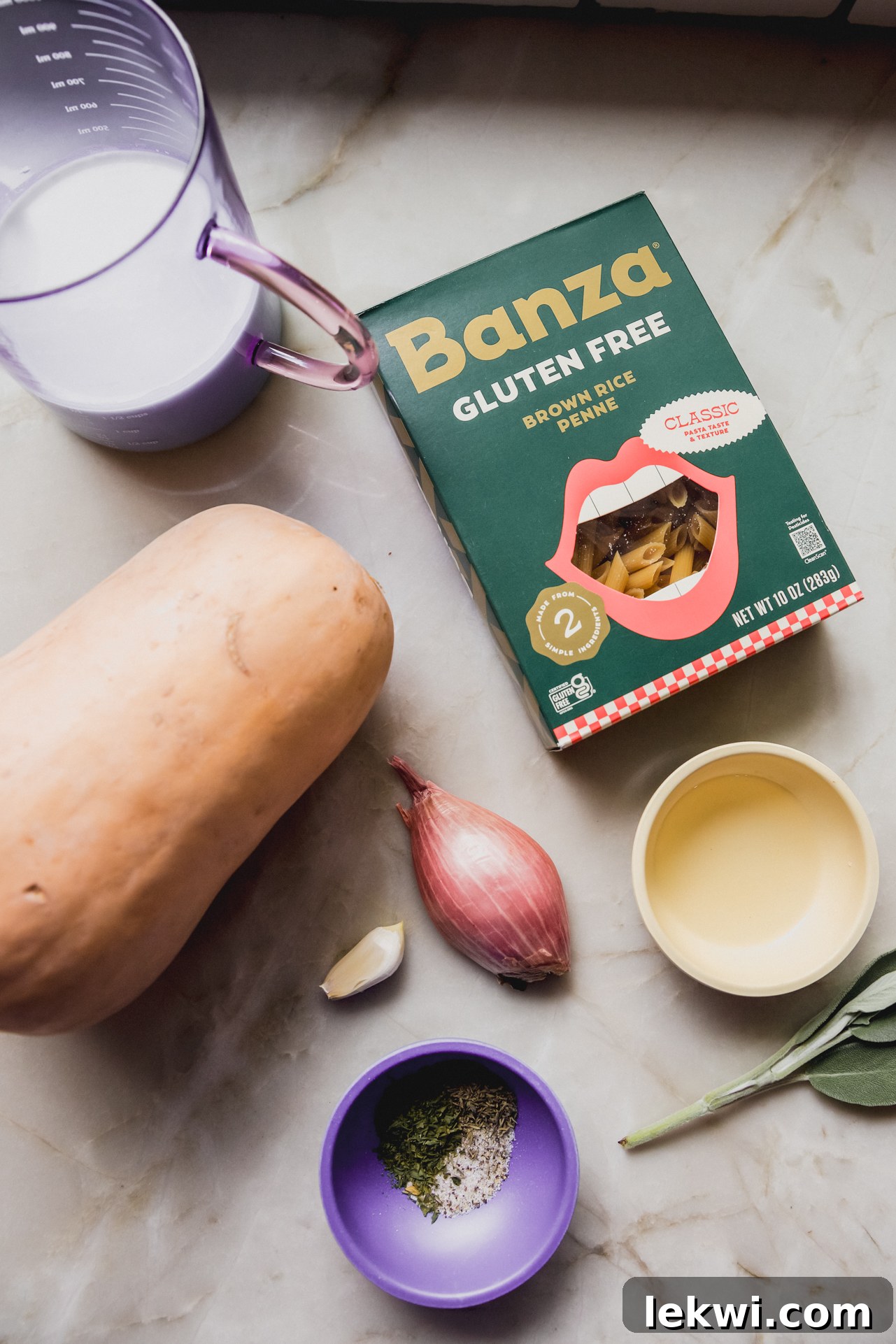 Various fresh ingredients for butternut pasta bake laid out in separate bowls, including cubed butternut squash, shallots, garlic, herbs, pasta, and cheese, on a light surface.