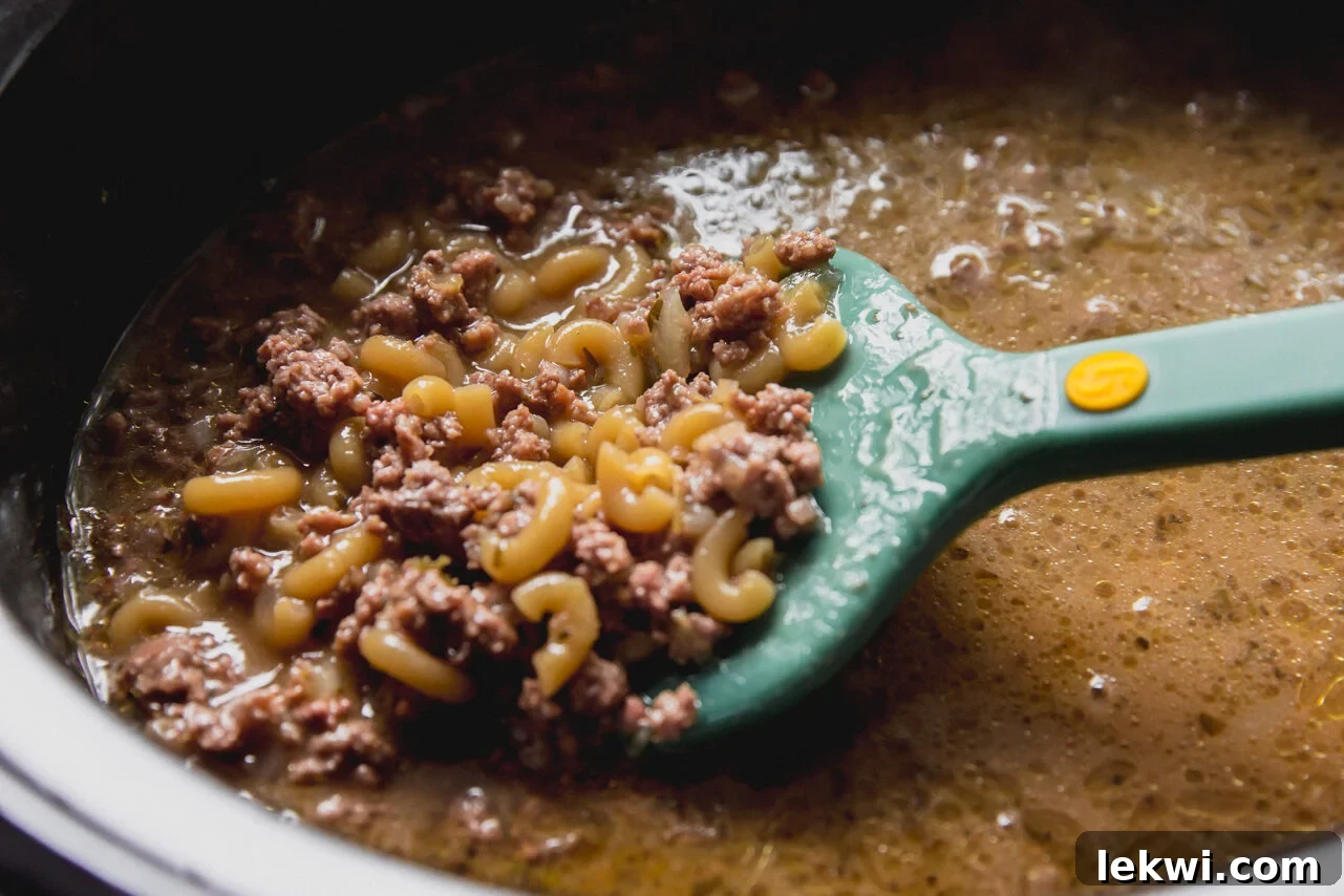 The slow cooker filled with ground beef, broth, pasta sauce, and almond milk, with dry pasta added on top, ready to be cooked.