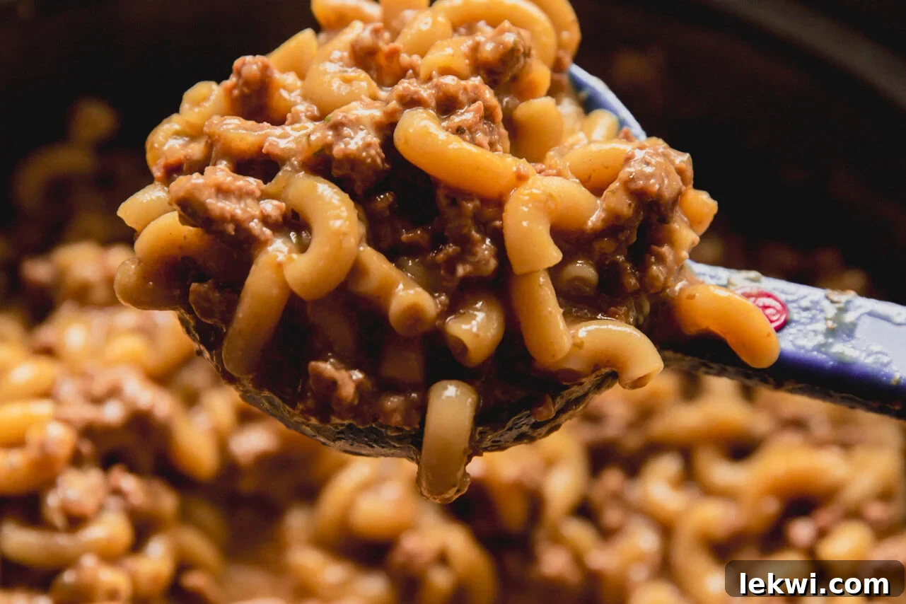 A serving spoon lifting a generous scoop of cheesy slow cooker hamburger helper from the pot.