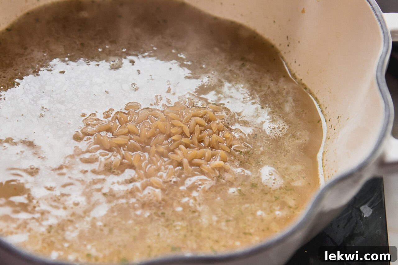 Orzo pasta cooking in a pot with chicken broth, garlic, and shallots, bubbling gently.