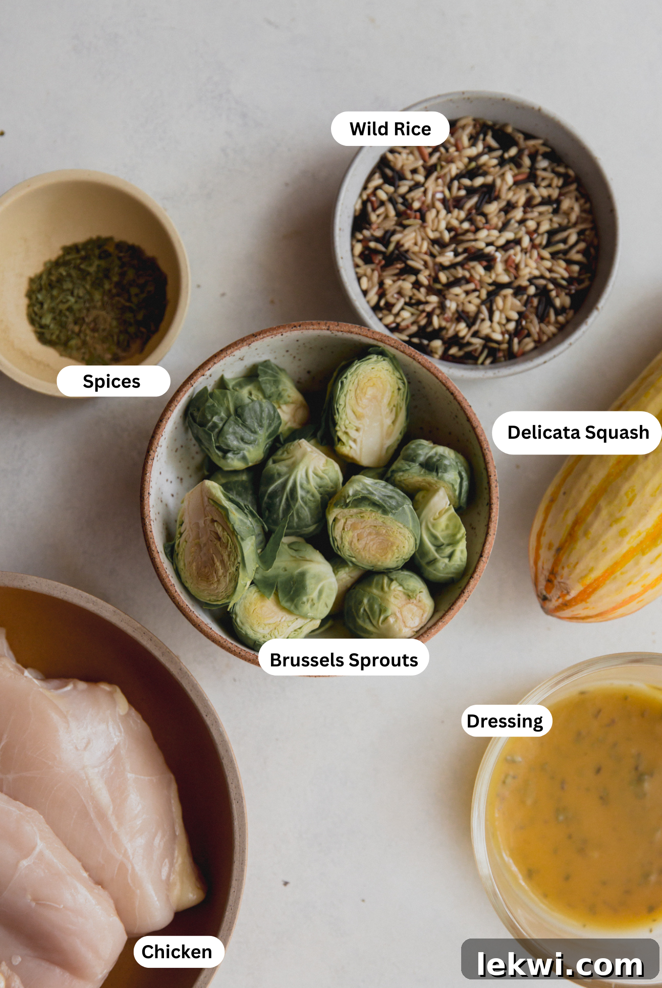 A spread of fresh ingredients laid out on a table: delicata squash, brussels sprouts, kale, chicken breast, wild rice, apples, cranberries, and dressing components for autumn squash and chicken harvest bowls.