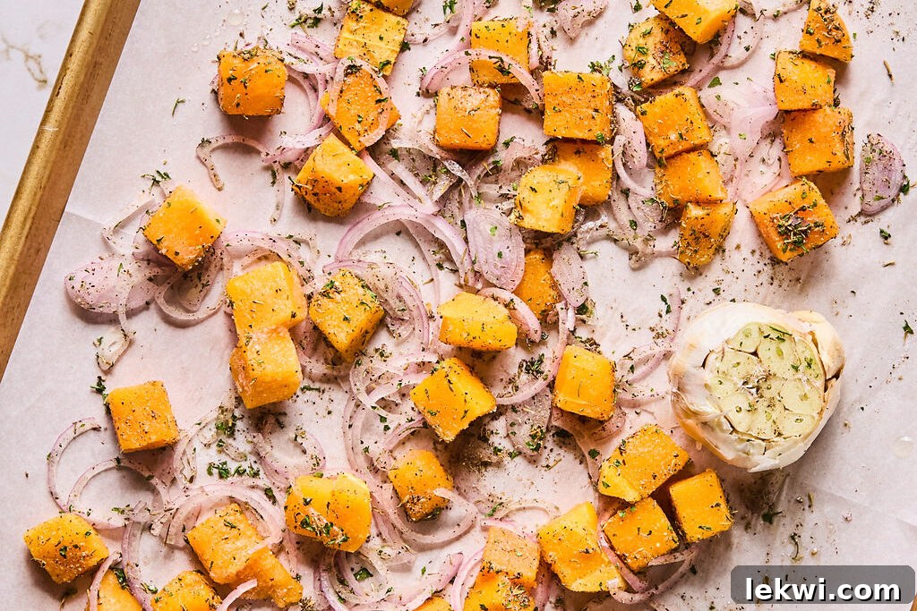 Butternut squash cubes, sliced shallots, and a head of garlic prepared for roasting on a baking sheet.