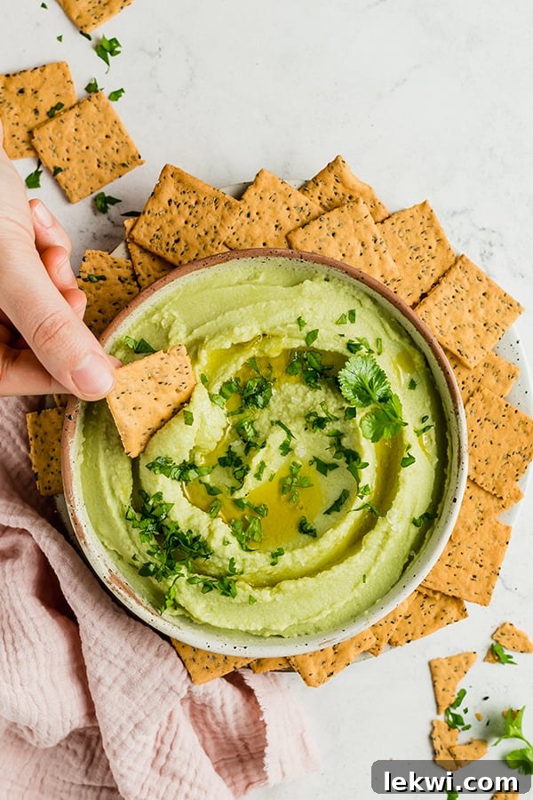 A cracker being dipped into a bowl of creamy avocado cauliflower hummus.