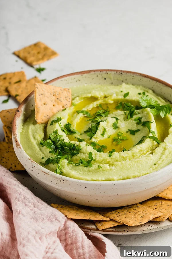 A cracker poised to dip into a bowl of perfectly green avocado cauliflower hummus.